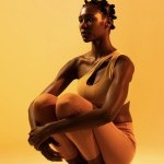 Dark skinned woman squatted wearing warm tone lathering wear with warm lighting resembling magic hour.