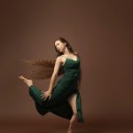 Female identifying dancer in green dress with right leg licked behind her in a front of a brown backdrop. 