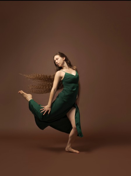 Female identifying dancer in green dress with right leg licked behind her in a front of a brown backdrop. 