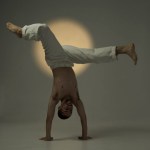 man on his hands, one leg in front of his body and the other bent behind him, in front of a warm light, while wearing white pant