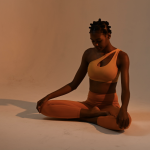 young black female yoga instructor sitting in a lotus pose wearing warm tone athletic wear