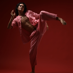 male dressed in pink pant suit wit leg leg lifted in front of a red backdrorp