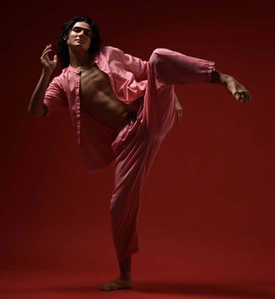 male dressed in pink pant suit wit leg leg lifted in front of a red backdrorp