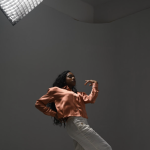 young lady moving wearing white pants and coral long sleeve shirt with lighting above 