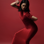 cis woman dressed in red dress with both legs bent in front of a red backdrop.