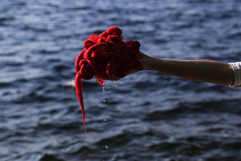 red yarn sculpture held by a hand in front of a body of water