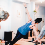 Three people in athletic wear are doing side planks on Pilates reformer machines in a well-lit studio. They are positioned in a 