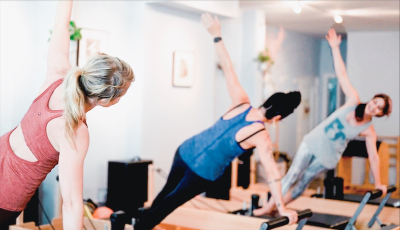 Three people in athletic wear are doing side planks on Pilates reformer machines in a well-lit studio. They are positioned in a 