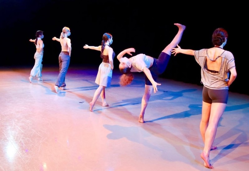 Five dancers stand in a line and one of them is in a back arch with their leg extended. 