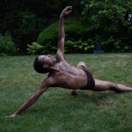 Male identifying dancer wearing black shorts extends his body while performing in the grass