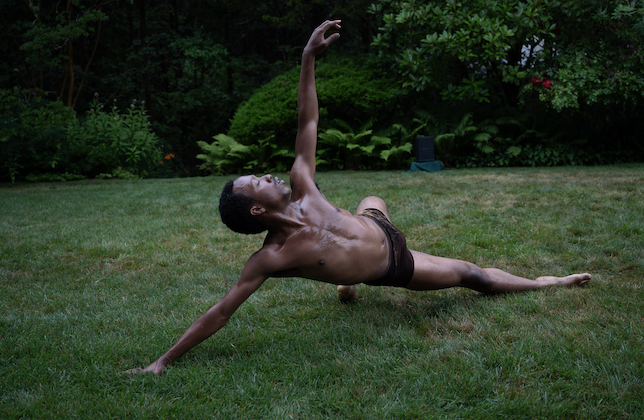 Male identifying dancer wearing black shorts extends his body while performing in the grass