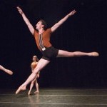 Male dancer jumping with legs & arms extended and a female dancer in the background