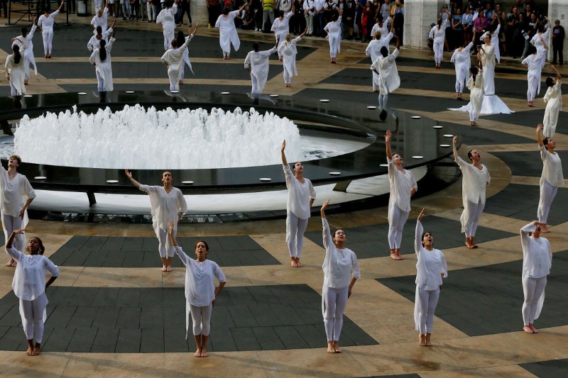 Dancers in white with one arm raised to the sky, encircling outdoor fountain on grey/beige granite plaza