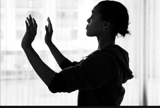 Dancer in silhouette with short hair and arms upraised palms facing ourward