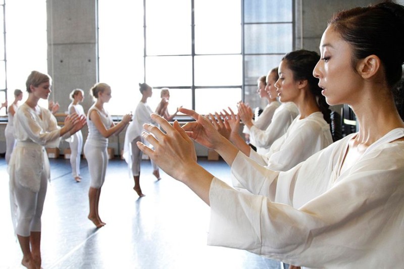 Dancers in white in lines arms raised at chest level, eyes looking down