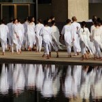 A large group of dancers in white cotton tops/tights walking away on outdoor plaza and reflected in the water of plaza's pool