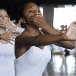 Dancers in white leotard with left hand over mouth and right hand reaching forward 