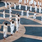 Dancers in white forming concentric circles on grey and beige outdoor plaza