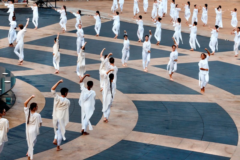 Dancers in white forming concentric circles on grey and beige outdoor plaza