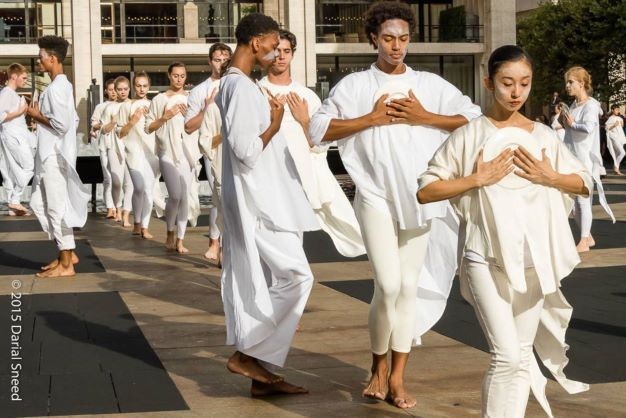 Outdoor procession of dancers on grey/tan plaza, dressed in flowing white cotton tops/tights, arms folded with plate on chest