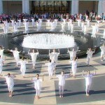 3 rows of dancers in white cotton tops and leggings on plaza encircling round fountain of water