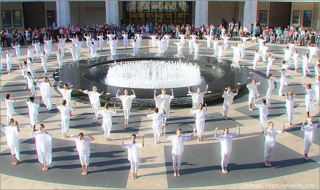 3 rows of dancers in white cotton tops and leggings on plaza encircling round fountain of water