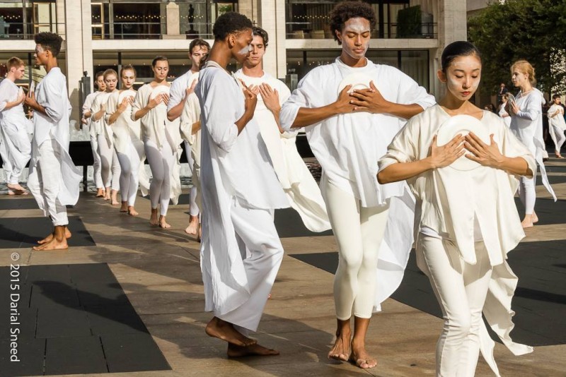 Processional of dancers in white tops and leggings on Plaza, hands resting on chest 