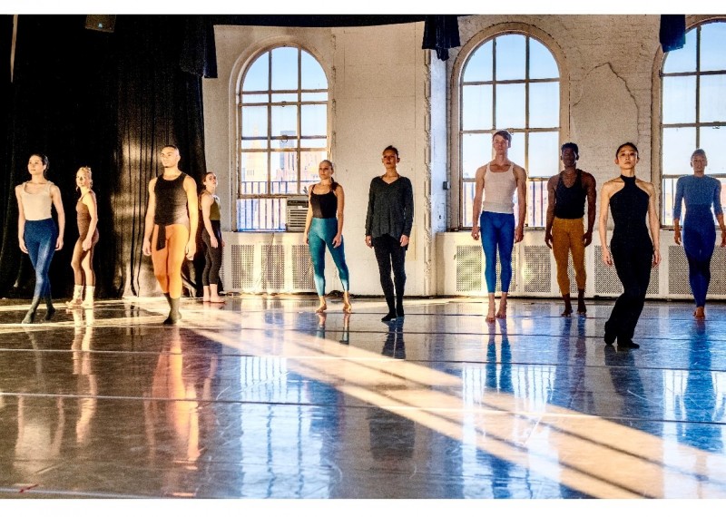 Multiple dancers facing forward in front of two windows with light beam on the floor 