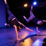 two dancers wearing black shirts and grey pants. they are both in one armed handstands.