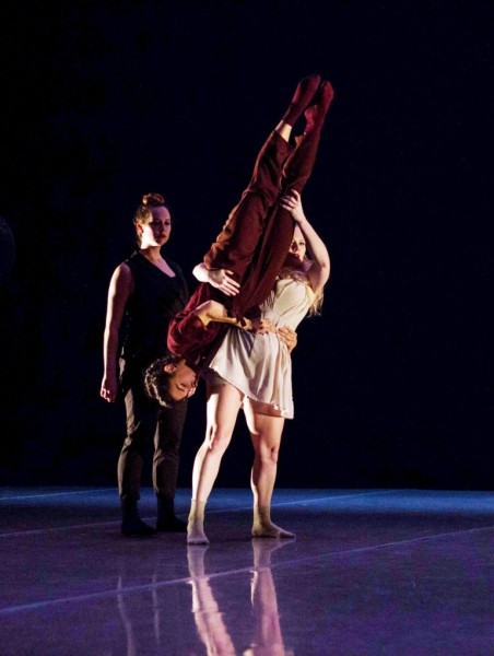 three dancers wearing black, red, and beige. one dancer is lifted upside down.