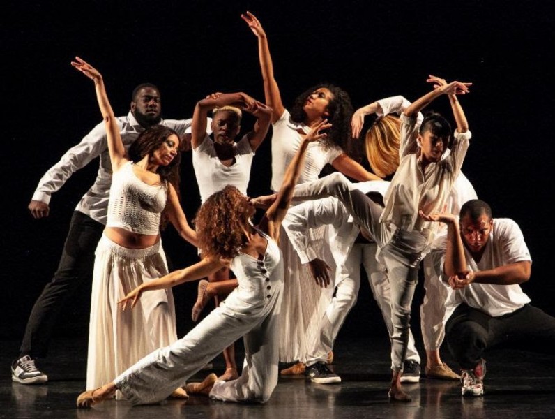 9 dancers dressed in white clothing dance together on theatrical stage. 