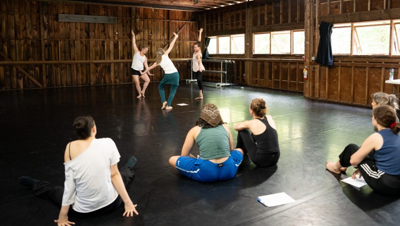 Three dancers move in a circle with one arm up and both knees bent. Five dancers watch along sitting on the floor.