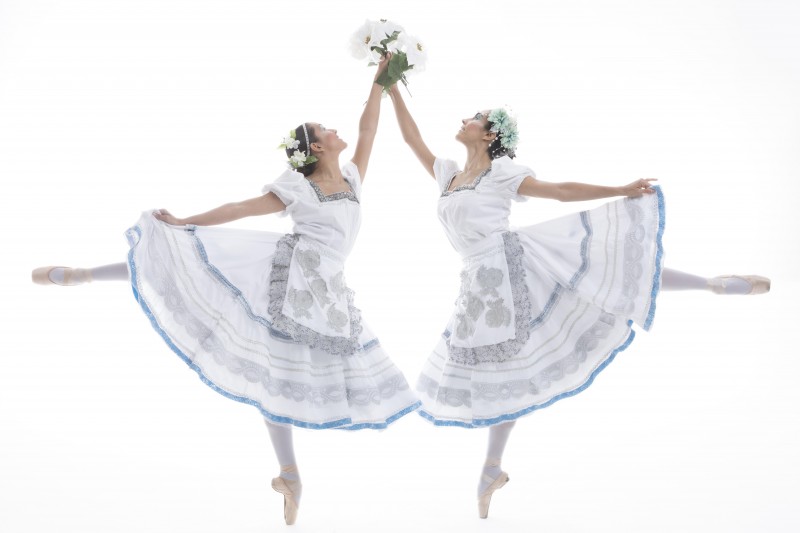 Two dancers wearing pointe shoes hold flowers and reach up, joining hands.