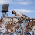 During a sunny day, sky bright light blue, a group of dancers wearing plastic bag costumes lift a female dancer on the air.