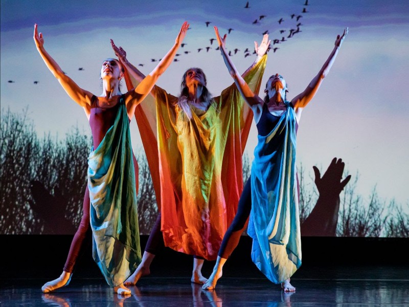 3 dancers in tunics. arms open to sky, image of sky and birds behnd them