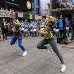 Two dancers side by side with one arm bent and one arm up straight, their head looking down, and one leg straight and one bent