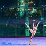 Dancer posed with her leg pointed up and her arms in the air, dancing in front of a city 