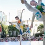 Image of two dancers in a white bodysuit with colour patches. The dancers are balancing on one leg and tilting to the left