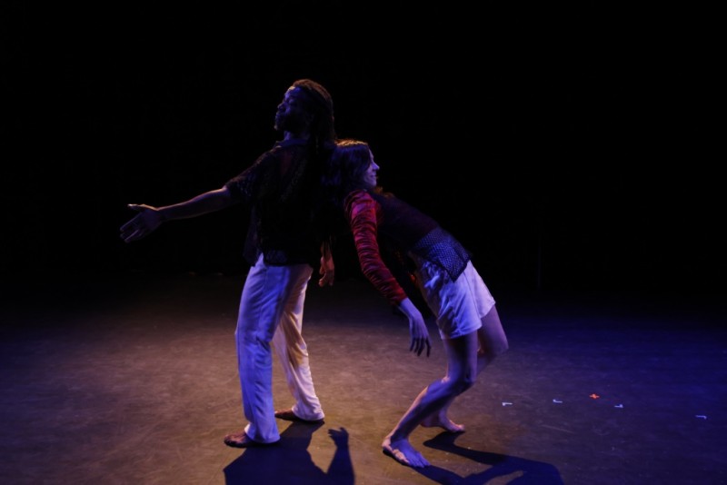 Two dancers alone on a stage lean on each other's backs.
