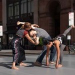 Two dancers extend forward and backward to embrace the other. Third dancer stands aside, leaning forward and extending leg back