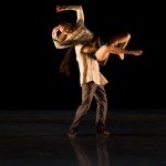 Dancer is lifted into the air, arms and legs bent. Side light illuminates dancer against black background