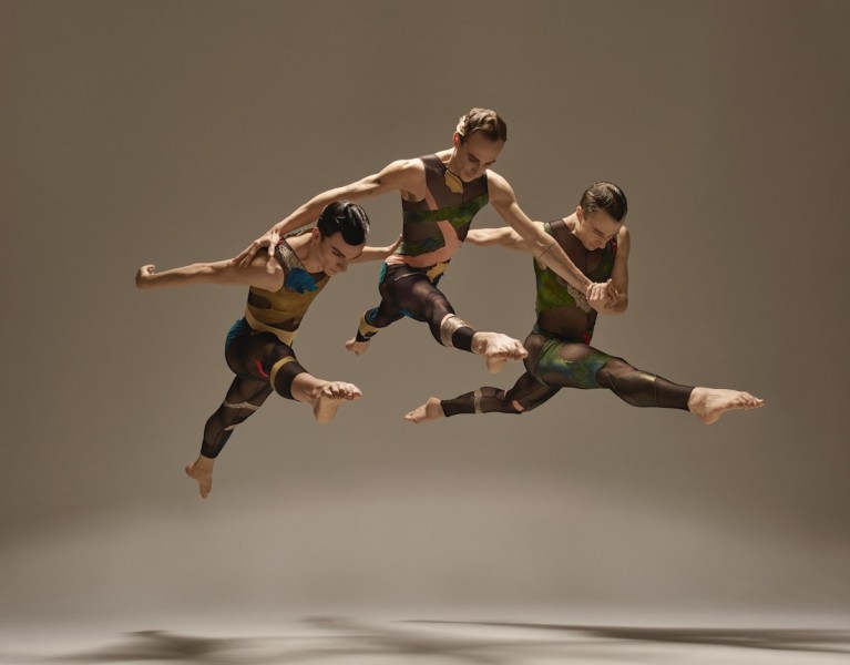 Three dancers leaping in the air with their arms linked wearing a brown costume with colored patches.