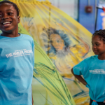 Two youth dancers stand in front of a colorful sheer backdrop, smiling