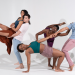 Five dancers wearing earth tones hold a group tableau.