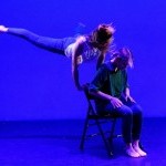 Dancer jumping behind chair holding back chair with dancer sitting in same chair bent forward