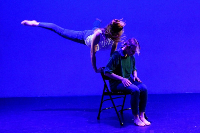 Dancer jumping behind chair holding back chair with dancer sitting in same chair bent forward
