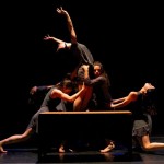 Dancers costumed in grey behind a wooden table with one dancer reaching up and the others all around her. 