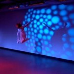 Dancer jumps in air with a background of colorful lighting projected on back wall