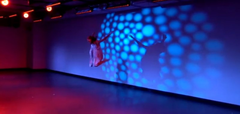 Dancer jumps in air with a background of colorful lighting projected on back wall