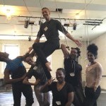 Six smiling people gathered around 1 person sitting in low trapeze, all dressed in dance clothing